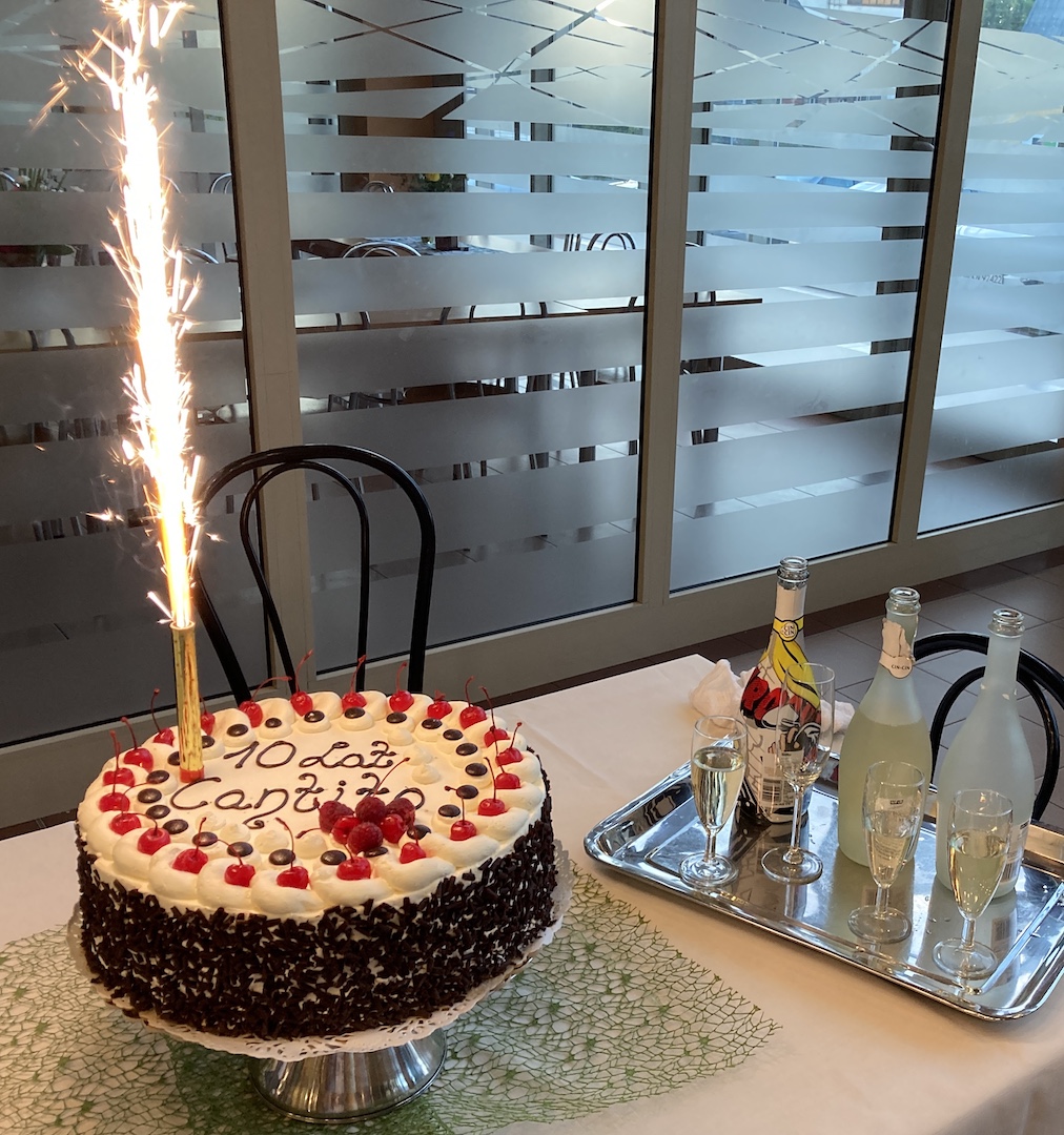 Na zdjęciu tort czekoladowy w białej polewie z owocami. Na torcie napis „10 lat Cantito” oraz płonąca raca. Obok na metalowej tacy otwarta butelka szampana i kilka kieliszków napełnionych szampanem.