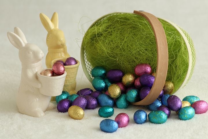 Grafika przedstawia koszyk wielkanocny, z którego wypadają czekoladowe jajka, a obok koszyka stoją dwa porcelanowe zajączki z koszykami w łapkach